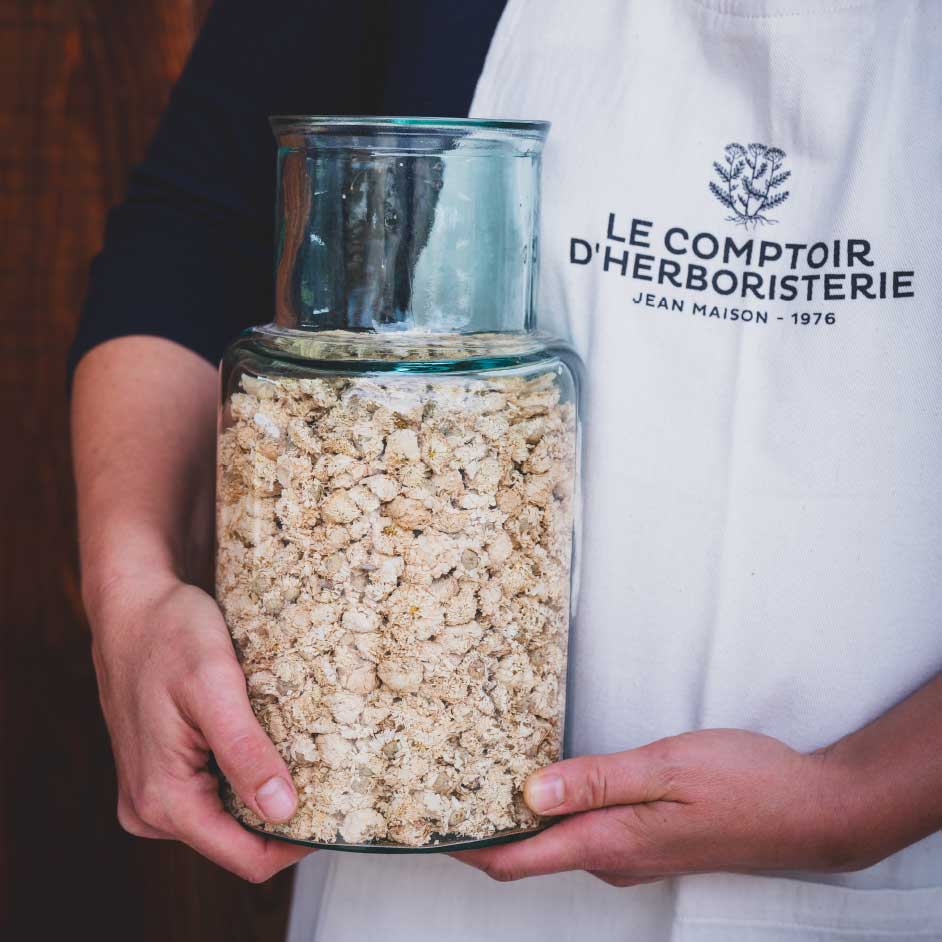 Tablier avec le logo. Personne tenant un grand pot en verre rempli de de fleur de camomille séchées. Photo Nadia Moléon